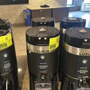 Fetco Luxus hot beverage dispenser. Features a single dispense valve, top handle, and an integrated drip tray base. Includes Starbucks branding.
