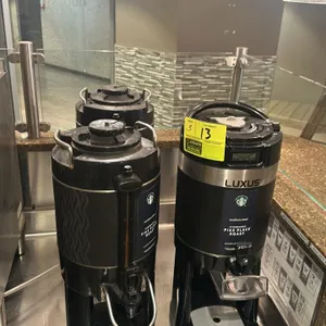 Assorted hot beverage dispensers. Includes (1) Fetco Luxus dispenser and (2) other black dispensers. Features spigots, carry handles, and drip trays.