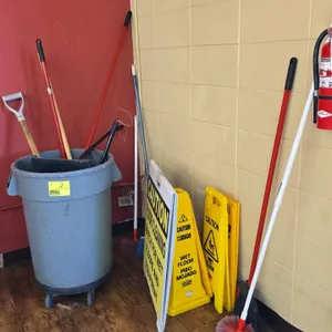 Package lot of janitorial supplies. Includes a gray plastic trash receptacle mounted on a caster dolly, various brooms, dusters, a dustpan, a squeegee