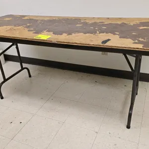 6'L folding table. Features a laminate top and a folding metal base. Note: Top surface exhibits heavy wear and peeling.
