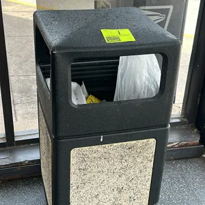 Poly outdoor waste receptacle. Features molded plastic construction in black with decorative aggregate-style side panels. Equipped with a hooded lid a