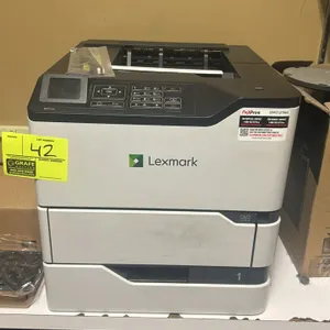 Lexmark MS725 monochrome laser printer. Features a digital display, push-button control panel, and front-loading paper drawer. Includes a packaged mai