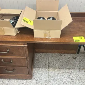 Wooden desk. Features a 3-drawer pedestal on the left with brass-tone decorative pulls, a wood-grain finish, and a raised rear backsplash border. 46"L