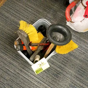 Assorted utility cleaning supplies. Includes a gray plastic bucket containing yellow-bristled scrub brushes, a plunger with a wooden handle, a hand sq