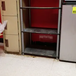4-tier open shelving unit featuring round gray metal corner posts and four dark shelves. Lockers, refrigerator, phone, and other background items are 