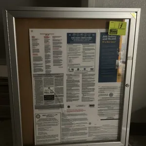 Enclosed cork bulletin board with aluminum frame. Features a single hinged glass door and cork interior. Currently contains labor law posters. Note: G