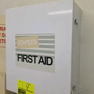 Staples metal wall-mounted first aid cabinet. Features white powder-coated metal construction, a single hinged door with dual latches, and two fixed i