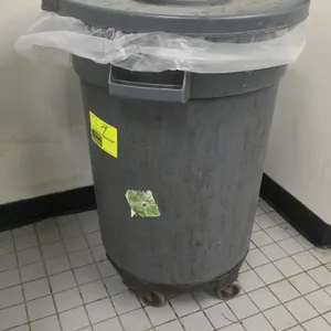 Grey plastic round trash can. Features a matching removable lid and dual molded-in handles. Mounted on a black circular twist-on dolly base with swive