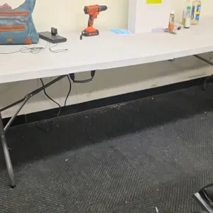 Poly folding table. Features a white plastic bi-fold tabletop that folds in half with an integrated center carrying handle. Mounted on a gray metal fo