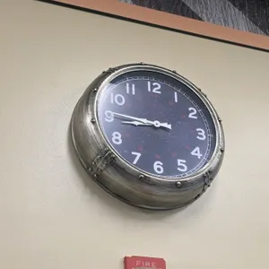 Naval design analog wall clock. Features a porthole-style metallic frame with rivet accents, a dark blue dial with white Arabic numerals, and white ha