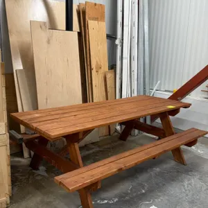 Wooden picnic table. Features a classic A-frame design with two attached benches. Constructed of wood with a brown stained finish. 8'L. Background ite