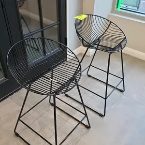 Metal bar height stool. Features a black wireframe design with a curved bucket seat and integrated footrest on a tapered base. 31"H.