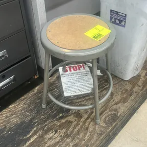 Metal utility shop stool. Features a gray-painted steel tubular frame, circular footring, and an inset round hardboard seat. Mounted on fixed legs wit