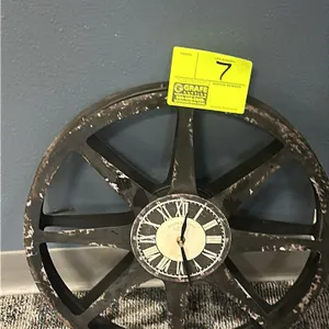Rustic wagon wheel style wall clock. Features a distressed dark finish with a spoke design, Roman numeral display, and black metal hands. Battery oper