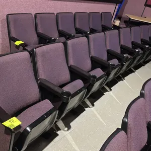 Movie theater seating row containing 4 connected seats. Features purple cloth cushioned foldable seats, black plastic outer back shells, and armrests 