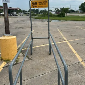 Metal shopping cart corral. Features tubular metal construction and an integrated yellow 'Please Return Carts Here' sign. 33"W x 134"L. Note: Backgrou