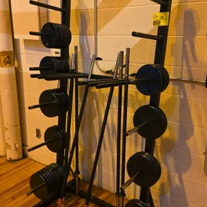 Weight storage rack with plates and bars. Features double-sided plate storage pegs, vertical bar holders, and steel frame construction. Includes an as