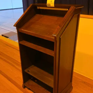 Portable wood podium. Features a slanted reading surface with an integrated light fixture, an upper open shelf, and two lower shelves with a cable pas
