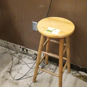 Wooden stool. Features a round seat, four-legged base, and dual-level support rungs. Natural wood finish.