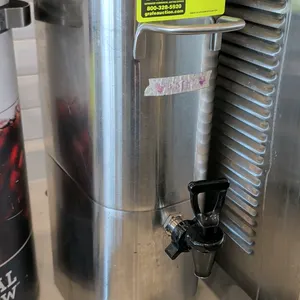 Stainless steel beverage dispenser. Features front lever dispensing faucet, front and rear carrying handles, removable black plastic lid, and drip tra