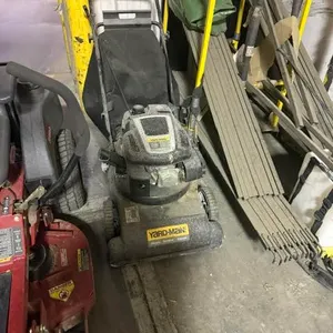 Yard-Man walk-behind lawn mower. Features a gas engine, black deck, rear bagger, and folding handle. Mounted on wheels. Note: Untested for functionali