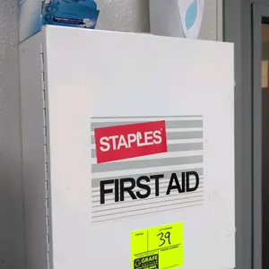 Staples metal first aid cabinet with contents. Features a white painted metal construction with 3 internal shelves, integrated door pouch organizer, a
