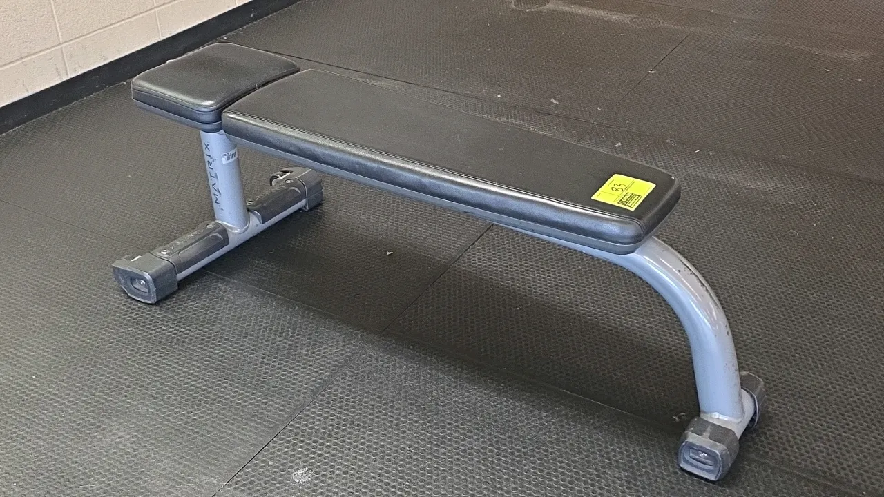 Matrix A59-03 flat exercise bench. Features black upholstered vinyl pads and a gray steel frame. Equipped with integrated transport wheels on the base
