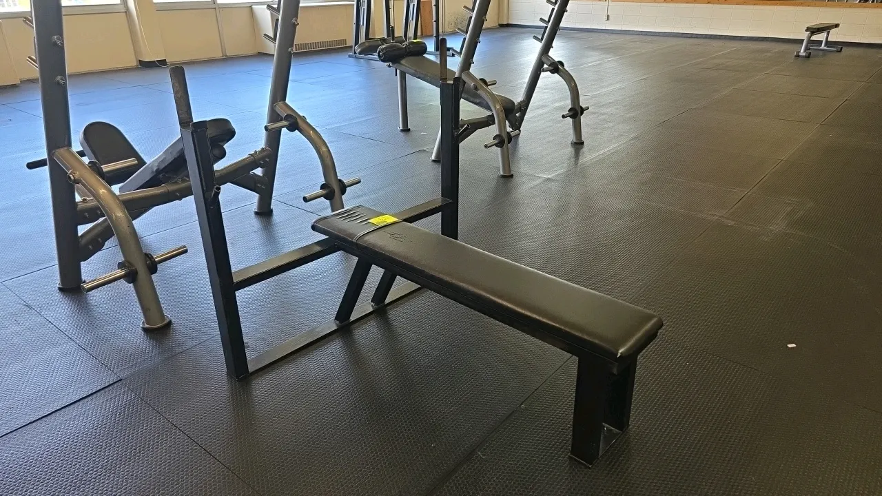 Olympic flat bench press. Features a black steel frame, dual-level barbell catches, and a black vinyl padded flat bench. Other gym equipment shown in