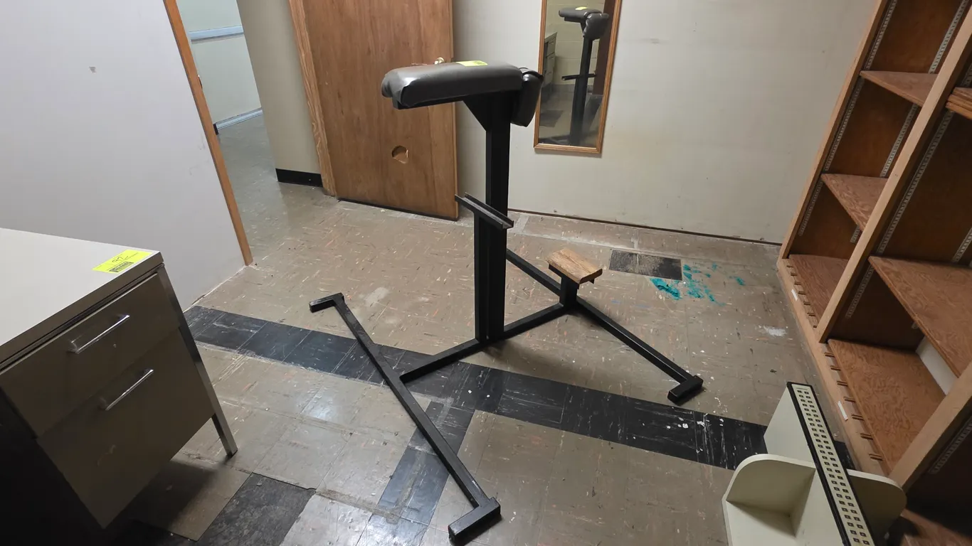 Metal donkey calf raise exercise station. Features a black square steel frame, a padded gray vinyl support cushion, and a wooden calf block. Includes