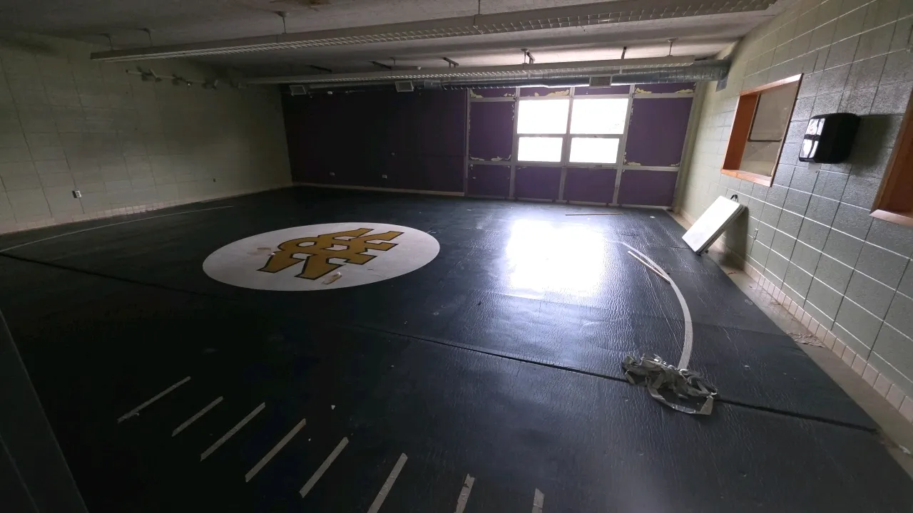 Room contents. Includes vinyl-surfaced wrestling floor mats and a plastic folding table. Mats feature a circular central logo. Note: Mats show tape re