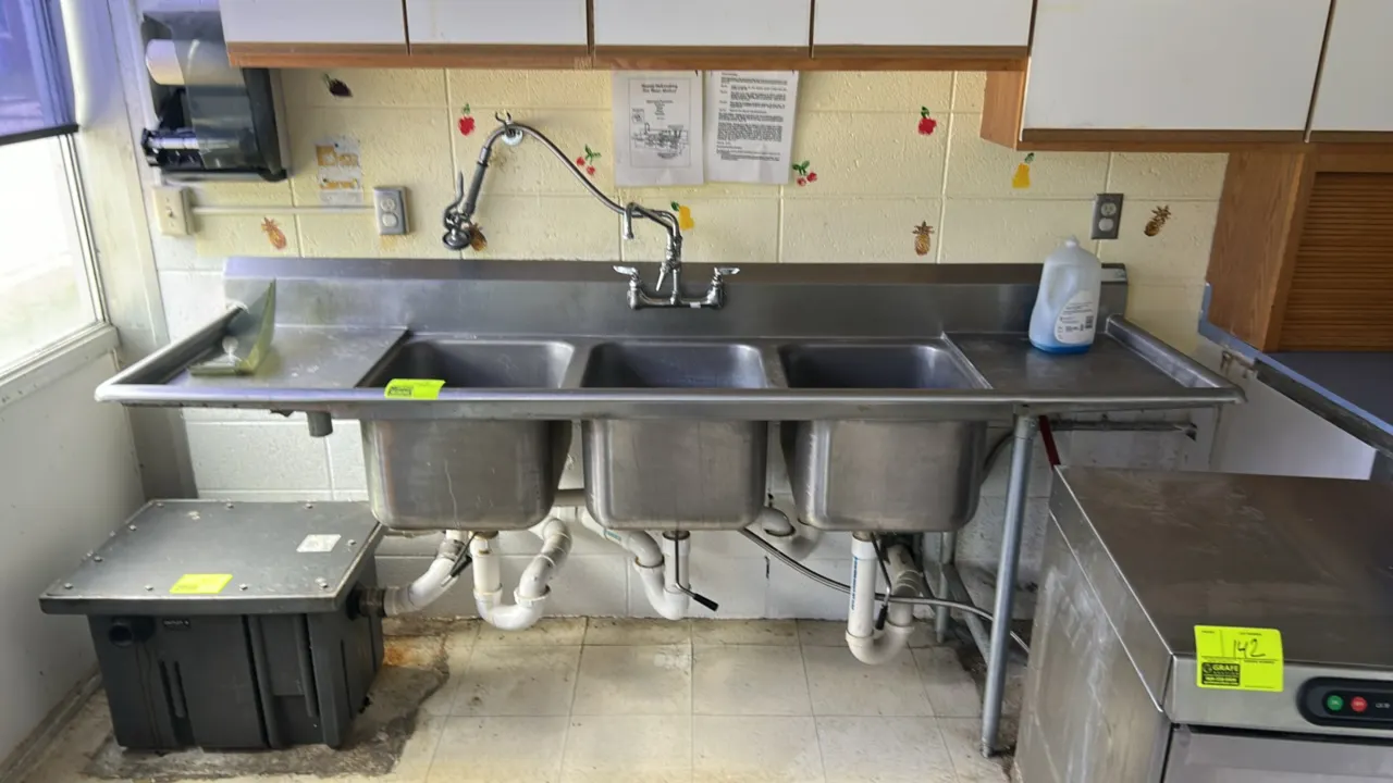 Stainless steel 3-compartment sink. Features left and right drainboards, a deck-mounted faucet with swing spout, and a wall-mounted pre-rinse sprayer.