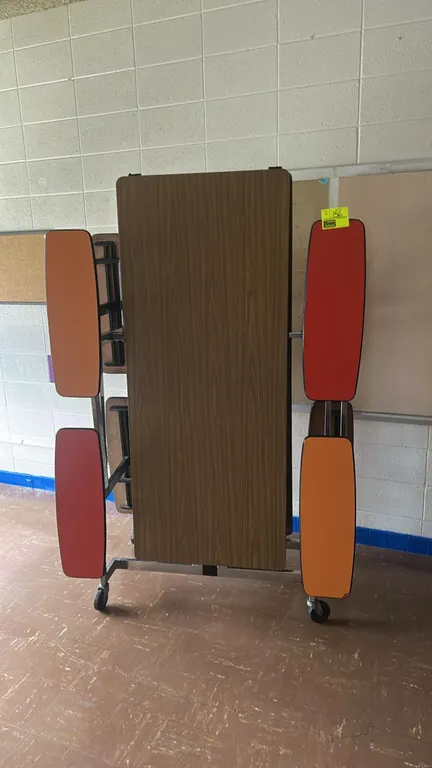 Foldable mobile cafeteria tables with attached bench seating. Features woodgrain laminate tops and colorful bench seats. Mounted on metal frames with