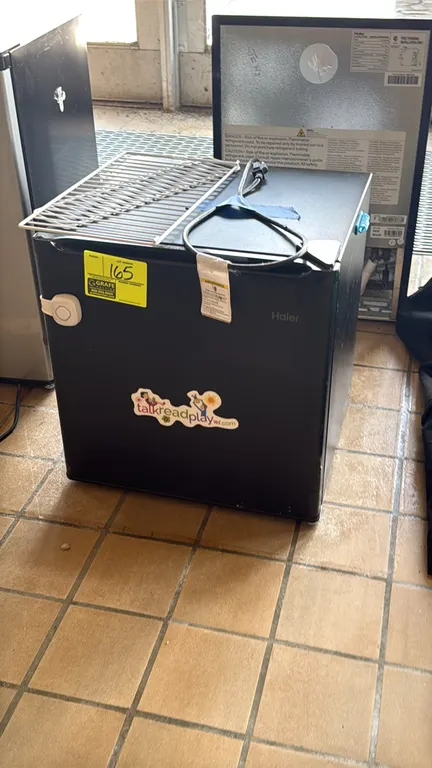 Haier countertop refrigerator. Features a black cabinet and single door. Includes one loose metal wire shelf. Note: Unit has a sticker on the front do