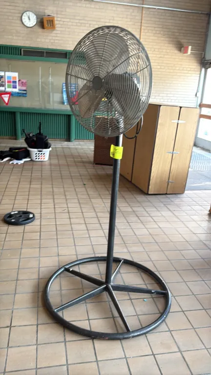 Pedestal fan. Features a 3-blade metal fan assembly enclosed in a protective wire safety cage. 22" diameter. Mounted on a metal pedestal pole with a c