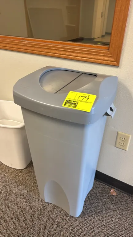 Rubbermaid Commercial Products poly trash can. Features gray plastic construction and a dual-flap swing lid. Note: Background items not included.
