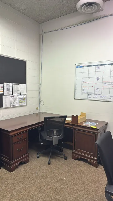 Wood corner desk with mesh-back office chair, dry erase board, and cork board. Desk features a brown wood finish, drawer pedestal, and cabinet storage