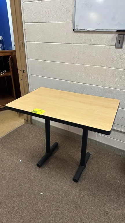 Laminate table. Features a light woodgrain laminate top with a black protective edge band. Mounted on a black metal T-base. Dimensions: 3'x2'.