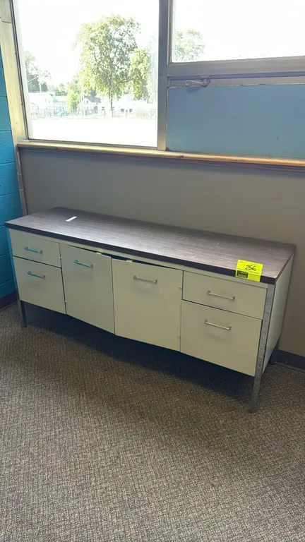 Metal credenza. Features a woodgrain-style laminate top, a central two-door storage cabinet with one shelf, and four drawers. 62"x18". Mounted on squa