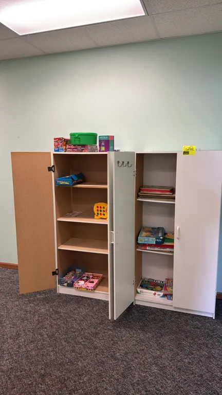 Laminate storage cabinets with various toys and games. Includes two white double-door cabinets featuring adjustable shelving and white laminate exteri