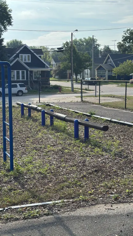 Metal balance beam. Features a dark-finished horizontal main pipe supported by six blue vertical posts. Designed for park or playground use. In-ground