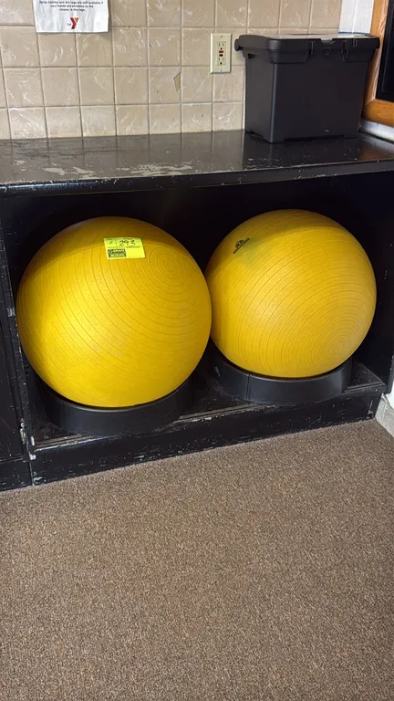 TheraBand exercise stability balls. Features yellow textured, slip-resistant design. Includes circular black plastic ring bases. Shelving and backgrou