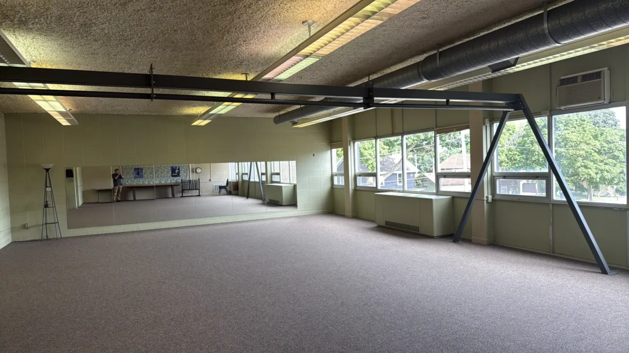 Contents of room. Includes a wood-grain conference table, a utility desk, (2) stacked upholstered chairs, a floor lamp, a wooden ballet barre, (2) wal