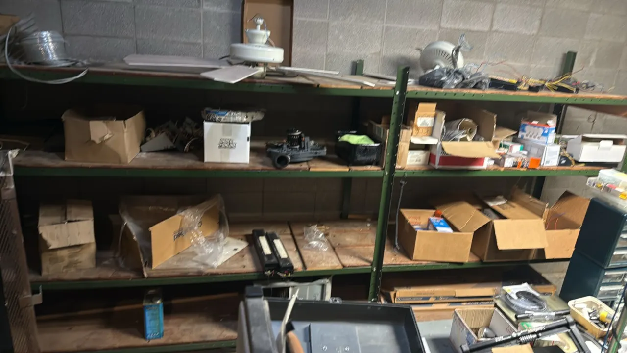 Metal 4-tier shelving rack with contents. Features green metal uprights and beams with wood deck shelving. Includes assorted electrical repair items,