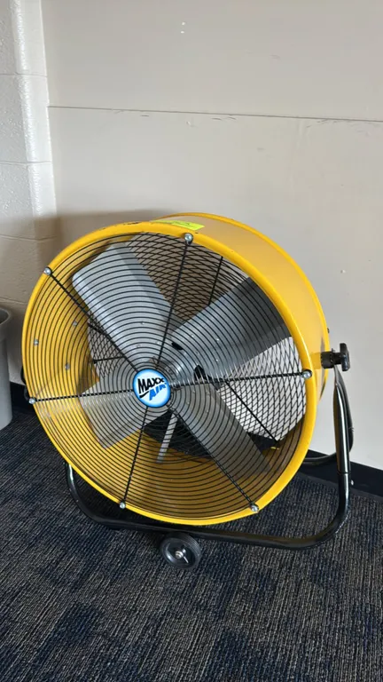MaxxAir metal floor fan. Features a yellow drum with black safety grilles, a 4-blade fan, and tilt-adjustable design. 24" diameter. Mounted on a black