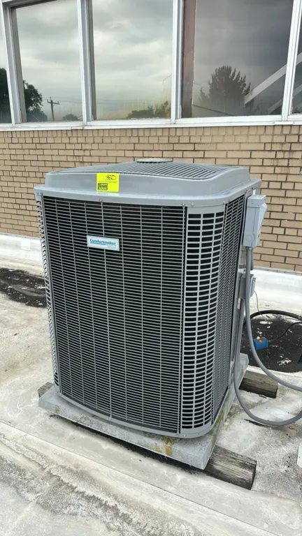 Comfortmaker SoftSound air conditioner condenser unit. Features top-discharge fan design with a protective metal outer grille. Mounted on a concrete p