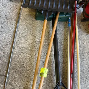 Assorted lawn and garden tools. Includes snow shovels with D-grip handles, a metal rake, brooms, a metal scraper rod, and miscellaneous utility handle