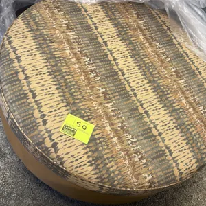 Round upholstered cloth ottoman. Features a patterned fabric top cushion in shades of tan, brown, and gray with a solid tan fabric-wrapped base. Mount