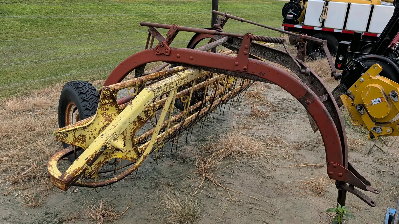 New Holland 56 side-delivery hay rake. Features ground-driven operation, tine basket assembly, and manual height adjustment cranks. Mounted on pneumat