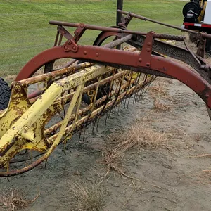 New Holland 56 side-delivery hay rake. Features ground-driven operation, tine basket assembly, and manual height adjustment cranks. Mounted on pneumat