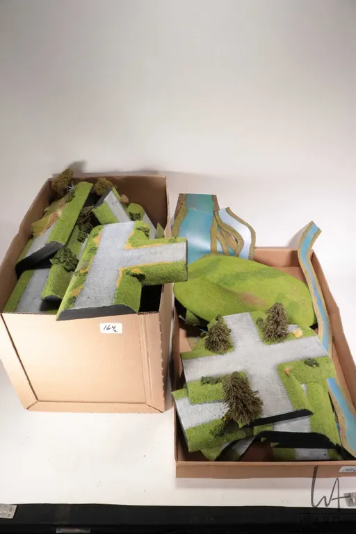 Two boxes containing a large selection of tabletop terrain including roads, rivers, grass mounts etc